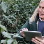 Elderly man engaging with a tablet in a lush, green outdoor setting.