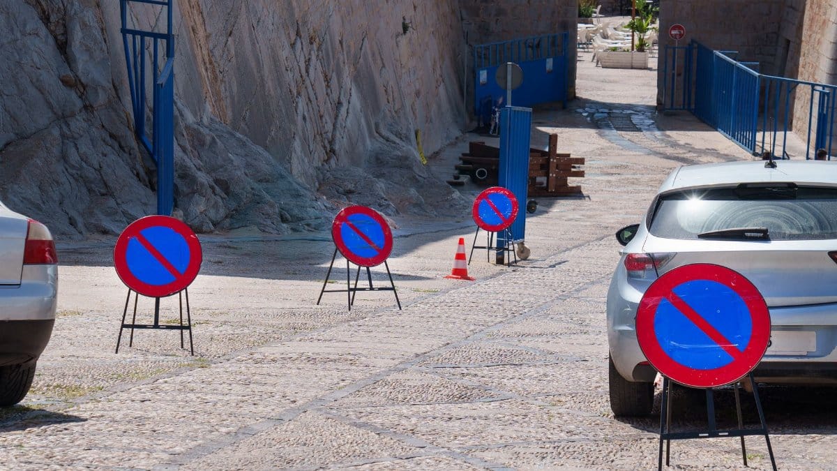 A row of no parking signs on a cobblestone street with parked cars, emphasizing road restrictions.