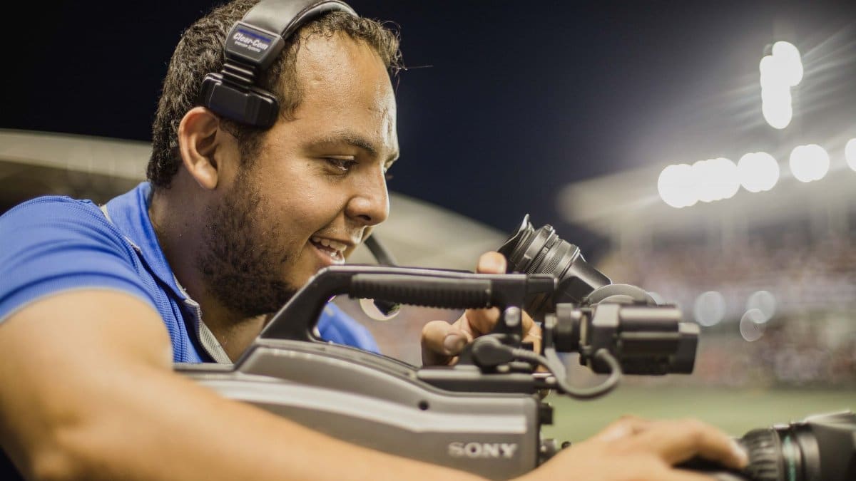 A cameraman with a headset operates a video camera outdoors at night, focusing on capturing footage.