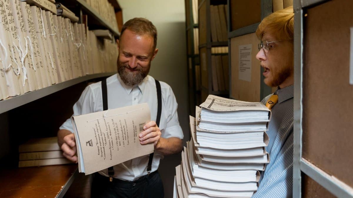 Two men sorting through stacks of documents in an archive room, showcasing teamwork and organization.