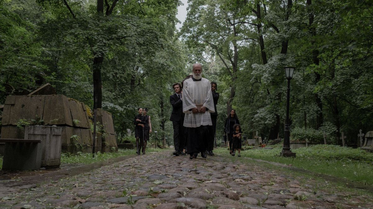 A group of mourners dressed in black walk through a peaceful cemetery path surrounded by lush trees.