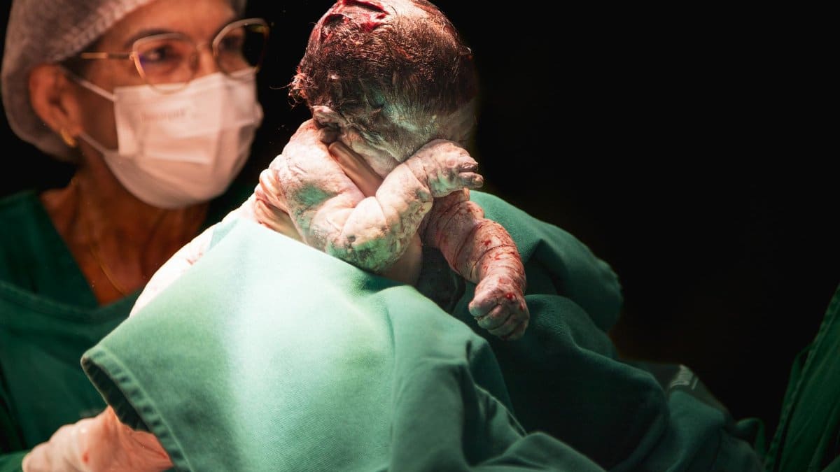 A newborn baby held by a nurse shortly after birth in a hospital setting.
