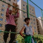 Two teenagers relaxing against a wire fence near urban apartment buildings on a sunny day.