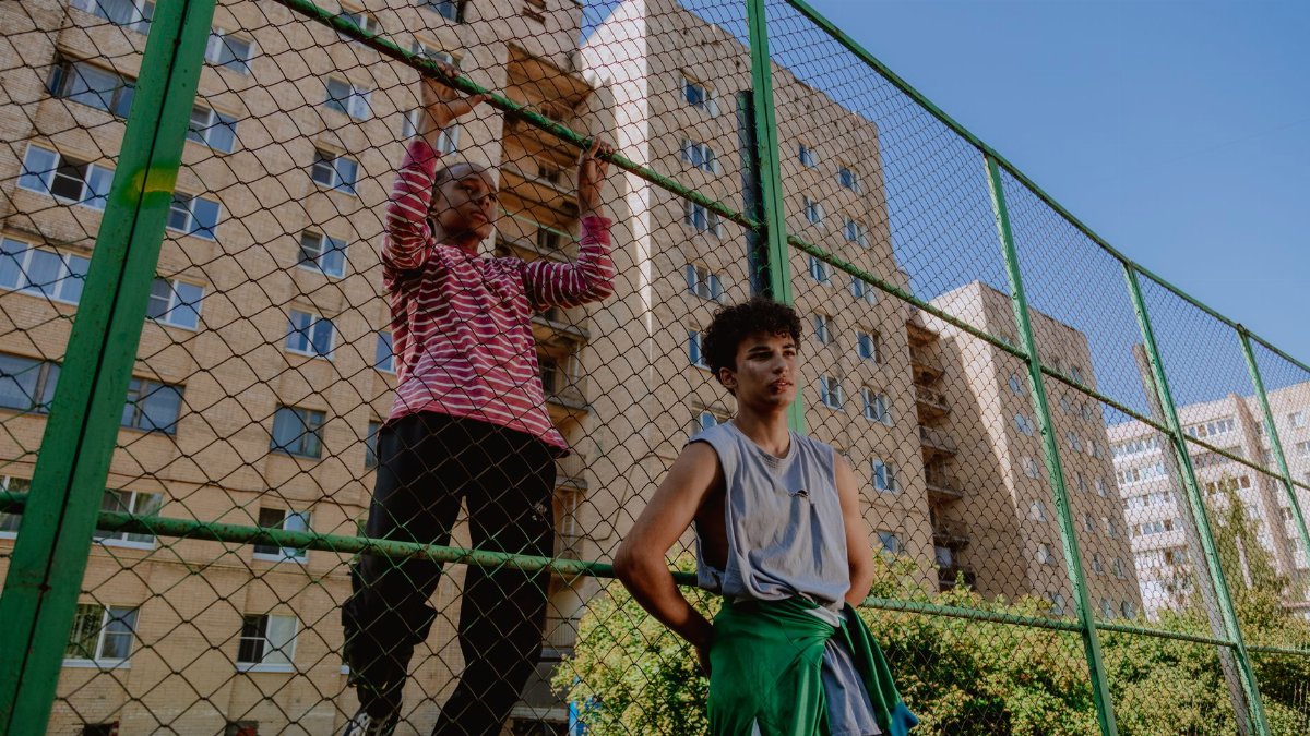 Two teenagers relaxing against a wire fence near urban apartment buildings on a sunny day.