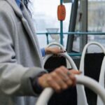 A person holding a rail inside a city bus, wearing a gray coat, with a street view outside.