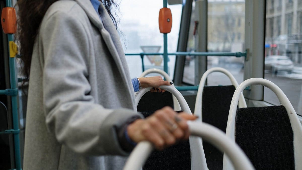 A person holding a rail inside a city bus, wearing a gray coat, with a street view outside.