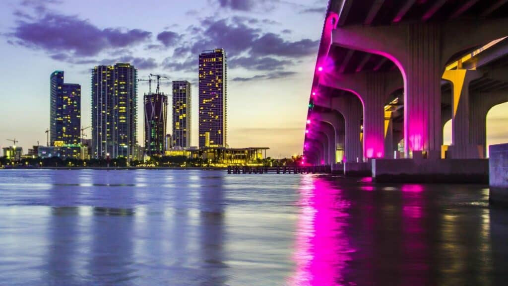 Captivating view of Miami skyline and illuminated bridge reflecting on water at dusk, vibrant city lights aglow.