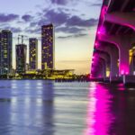 Captivating view of Miami skyline and illuminated bridge reflecting on water at dusk, vibrant city lights aglow.