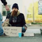A homeless man sits with a 'Will Work for Food' sign beside a shopping cart.