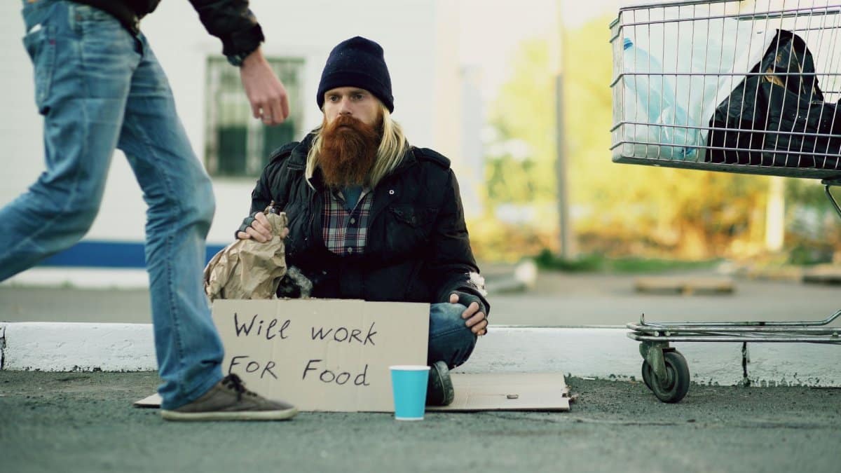 A homeless man sits with a 'Will Work for Food' sign beside a shopping cart.
