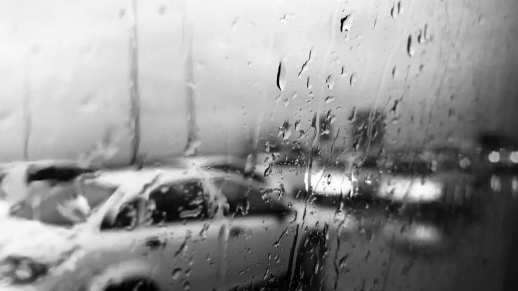 A black and white image capturing a car seen through rain-speckled glass.