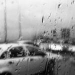 A black and white image capturing a car seen through rain-speckled glass.