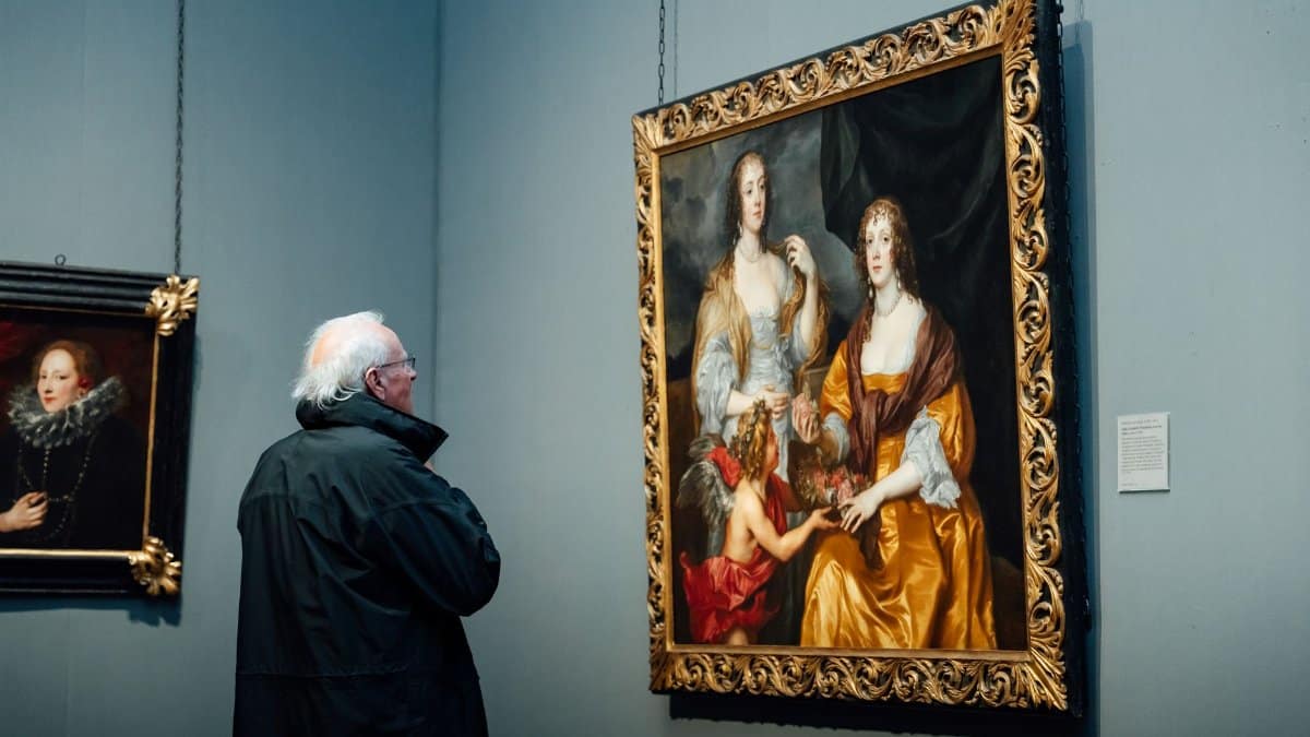 A senior man observes classical paintings in an art museum gallery.