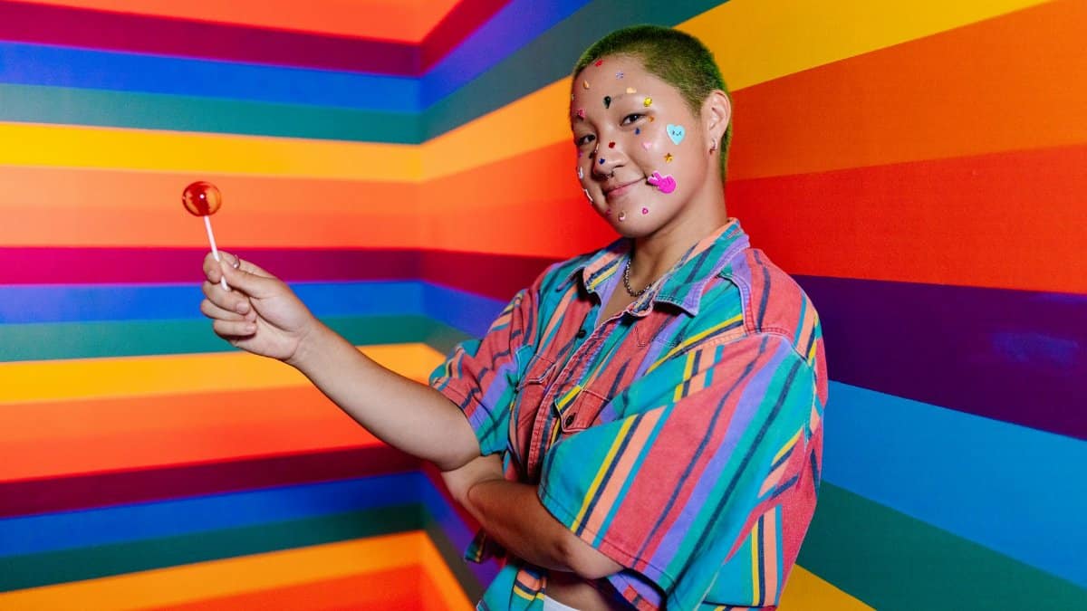 An adult poses playfully in a rainbow-colored room, holding a lollipop, with fun face decals.