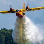 A firefighting aircraft releasing water to combat a forest fire in daylight.