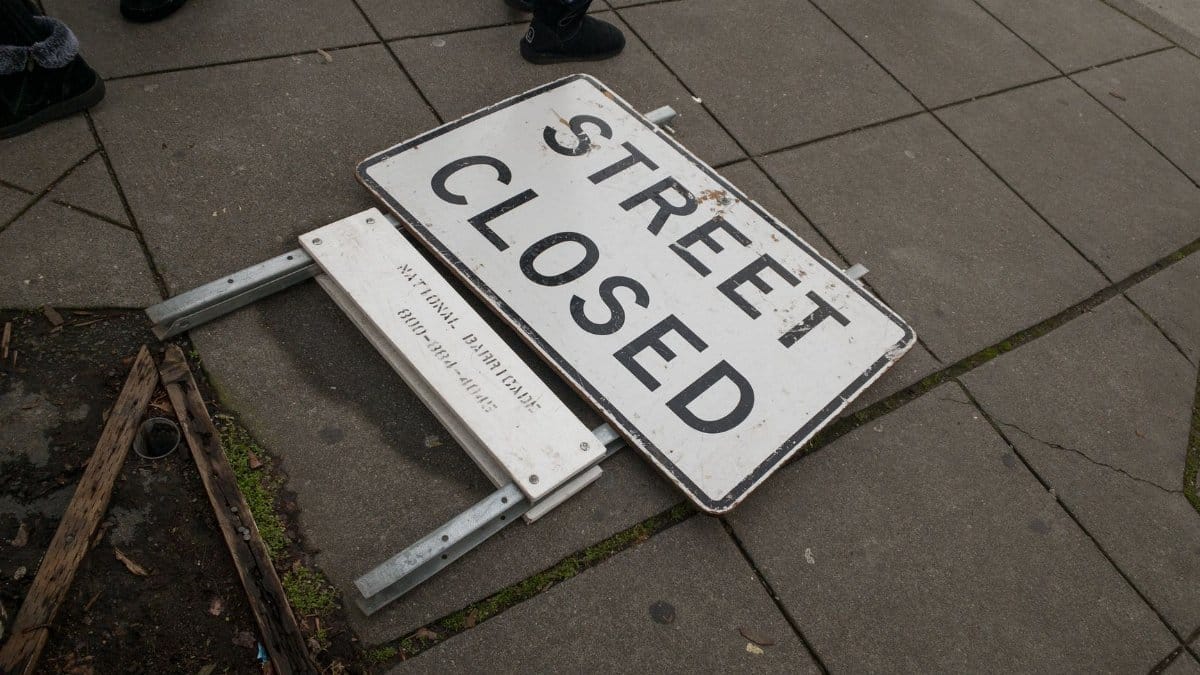 A fallen street closed sign on pavement with pedestrians walking nearby.