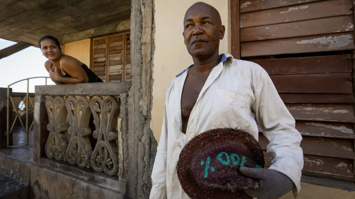 A man in a white shirt and glove holds a hat outside a rustic house as a woman observes from the balcony.