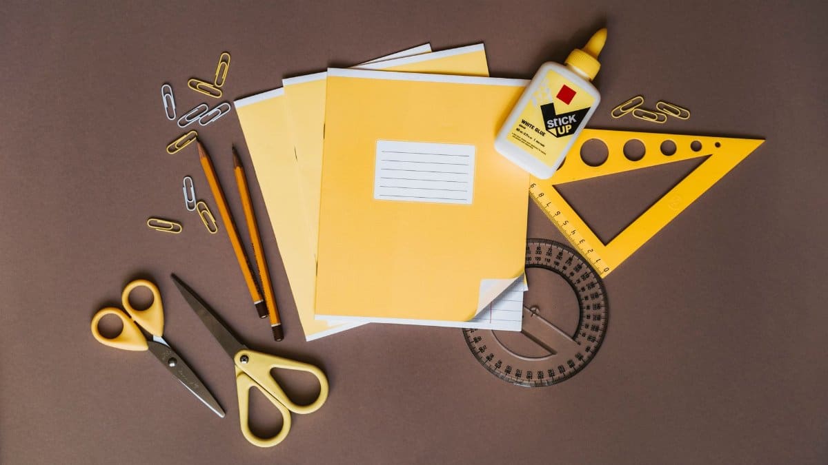 A flat lay of yellow stationery tools like pencils, scissors, and glue on a brown surface.