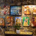 A vivid indoor display of animal and landscape paintings on a textured stone wall.