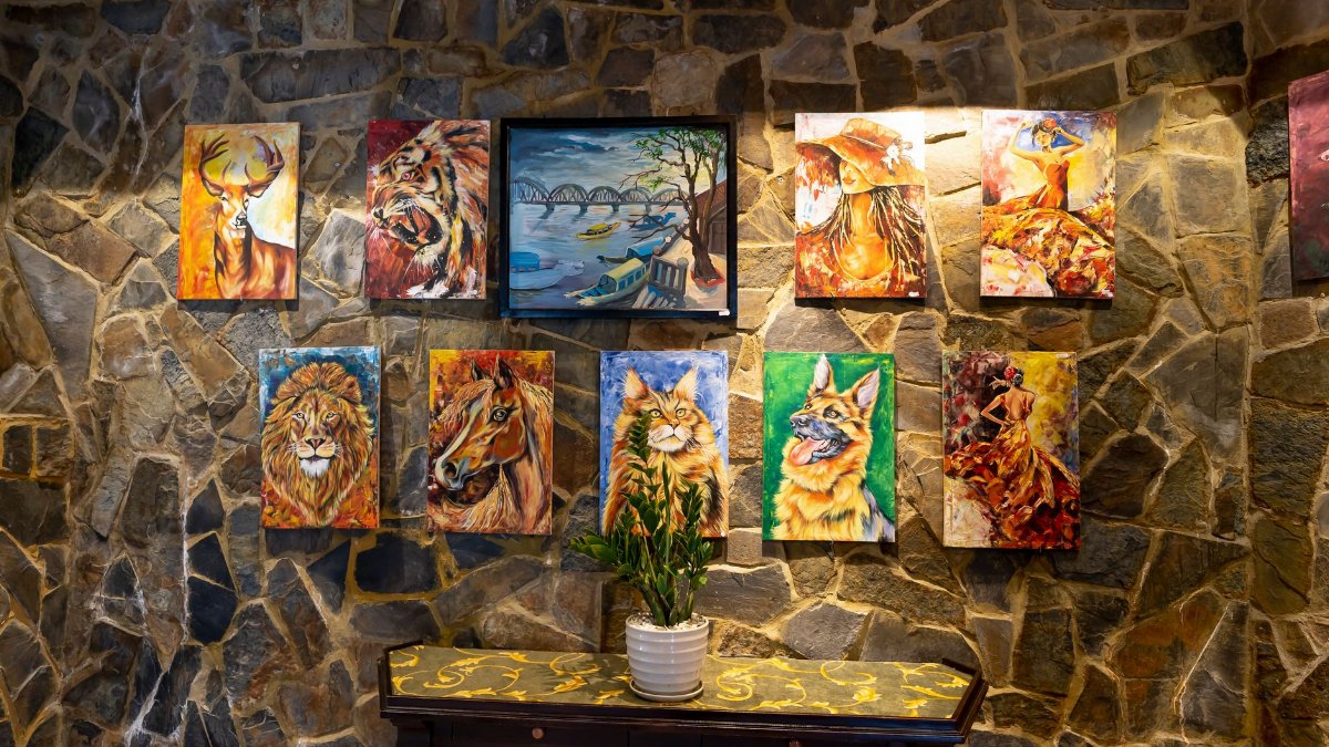 A vivid indoor display of animal and landscape paintings on a textured stone wall.