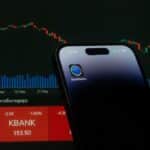 A mobile phone displaying a trading app in front of a financial market chart screen.