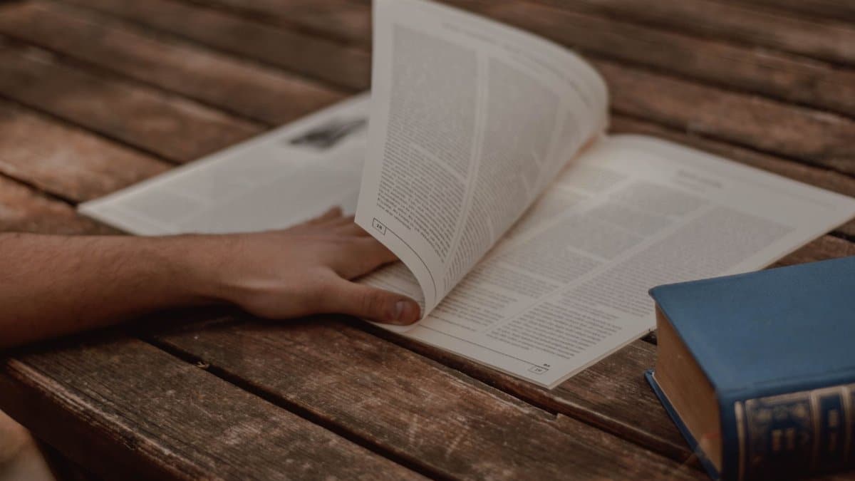 From above of crop unrecognizable male reading magazine with text in columns near old book