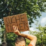 A man in a crowd holds a sign reading 'Suche Tickets' at an outdoor event, seeking extra admissions.