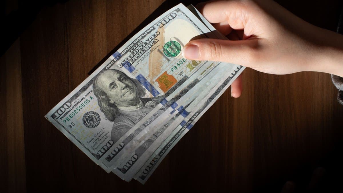 A hand holding a stack of 100 dollar bills on a wooden table, highlighting currency wealth.