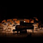 A dimly lit concert hall stage featuring an empty grand piano and chairs for an orchestra.