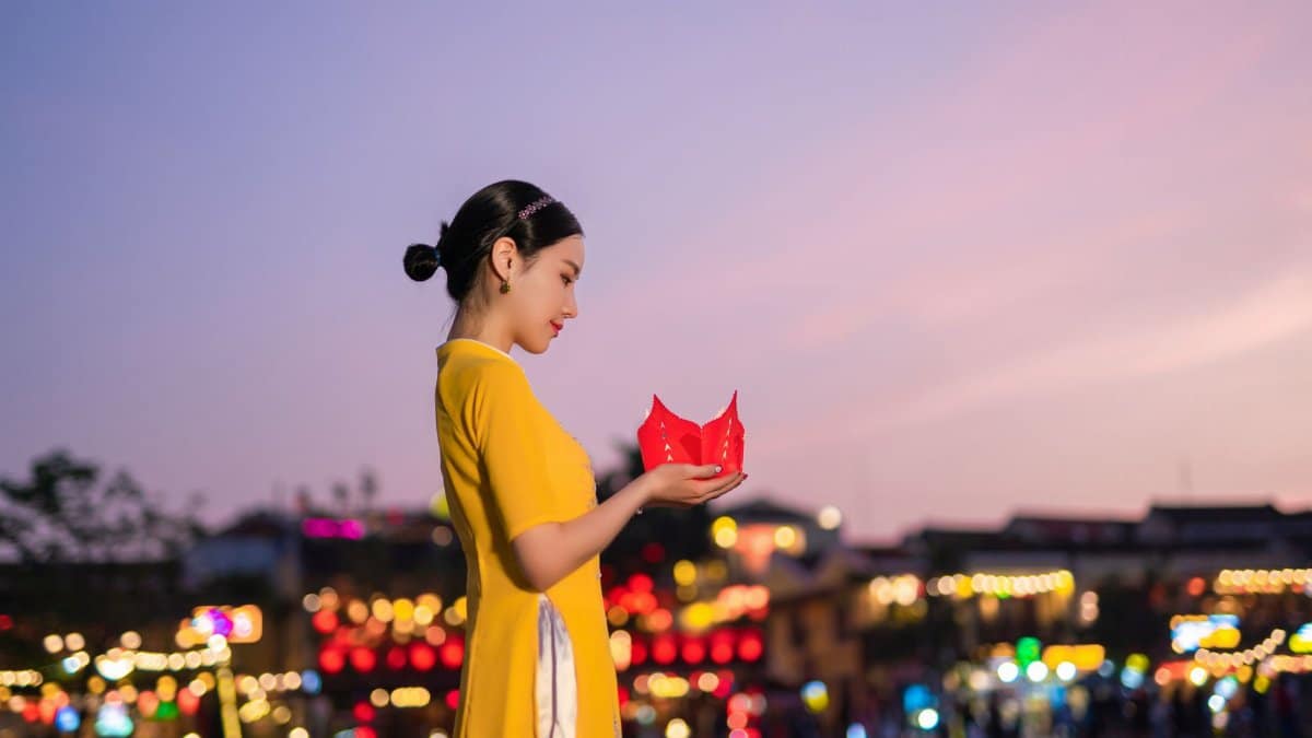 An Asian woman in a yellow dress holds a lantern amidst a vibrant, festive setting at twilight.
