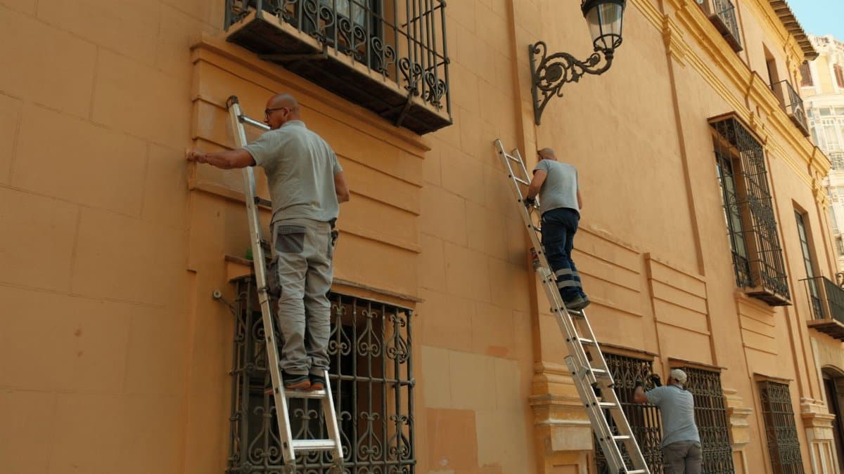 Three men on ladders working on the facade of a historic building in Spain, showcasing restoration efforts.