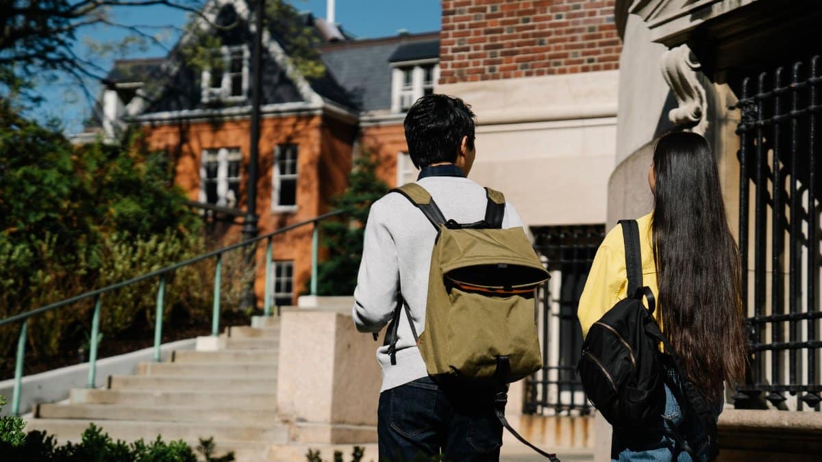 Two students with backpacks walking up stairs towards a brick building on campus.