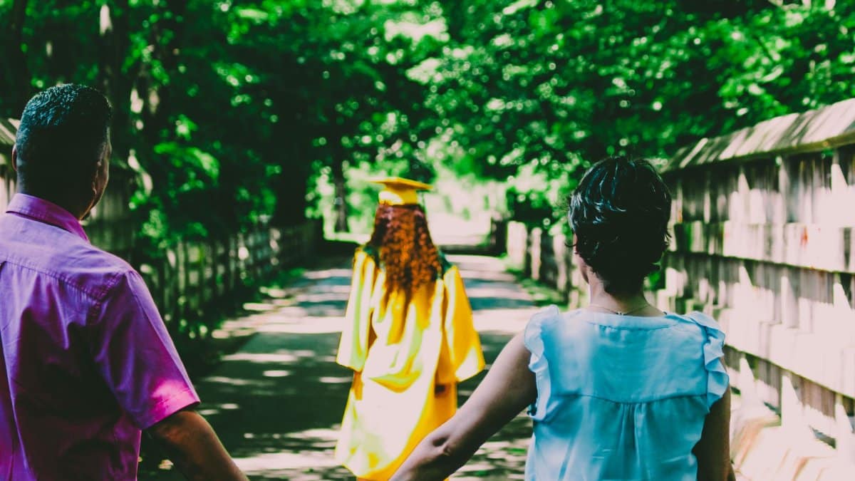 A graduate in a yellow gown walks ahead as parents hold hands outdoors, celebrating a new chapter.