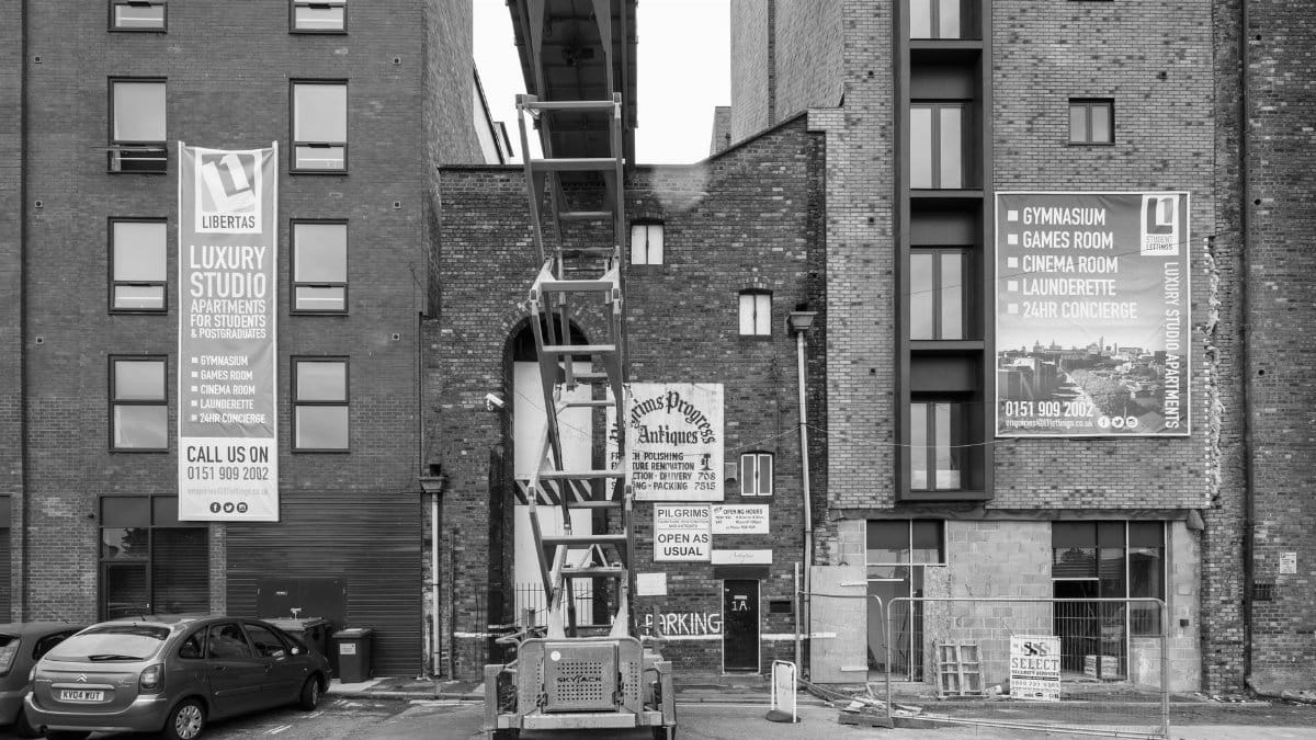 Black and white view of a construction site amidst urban buildings featuring banners and parked cars.