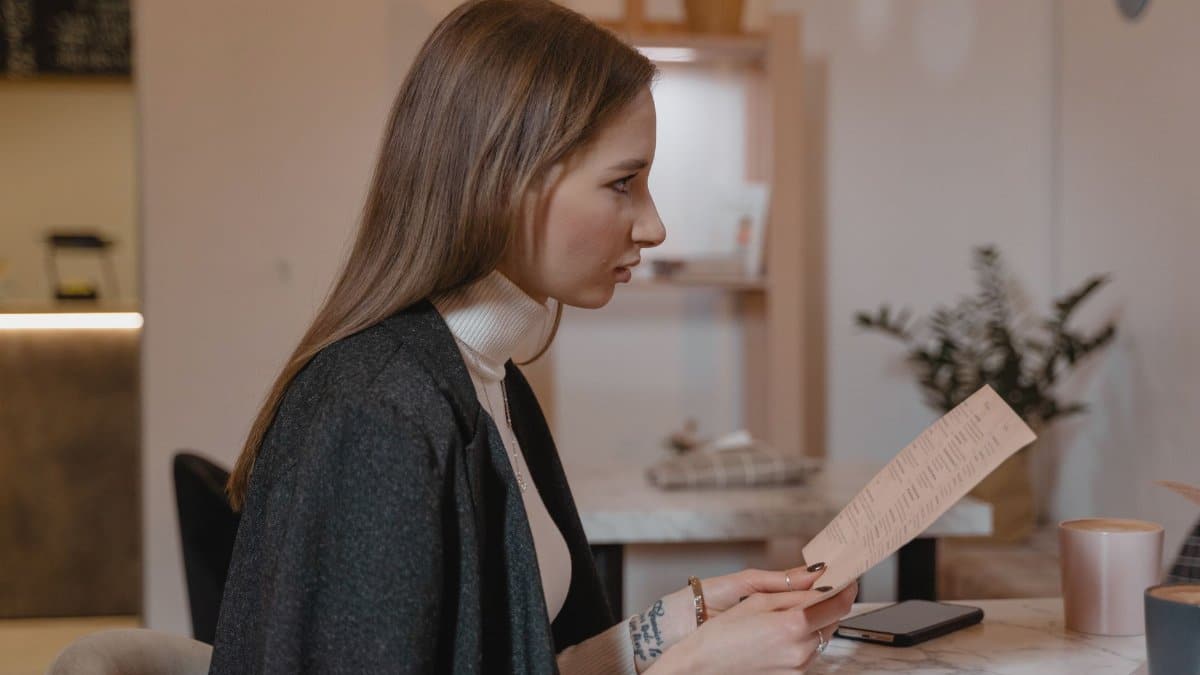 Side view of woman holding menu in a cozy café setting, wearing a turtleneck and jacket.