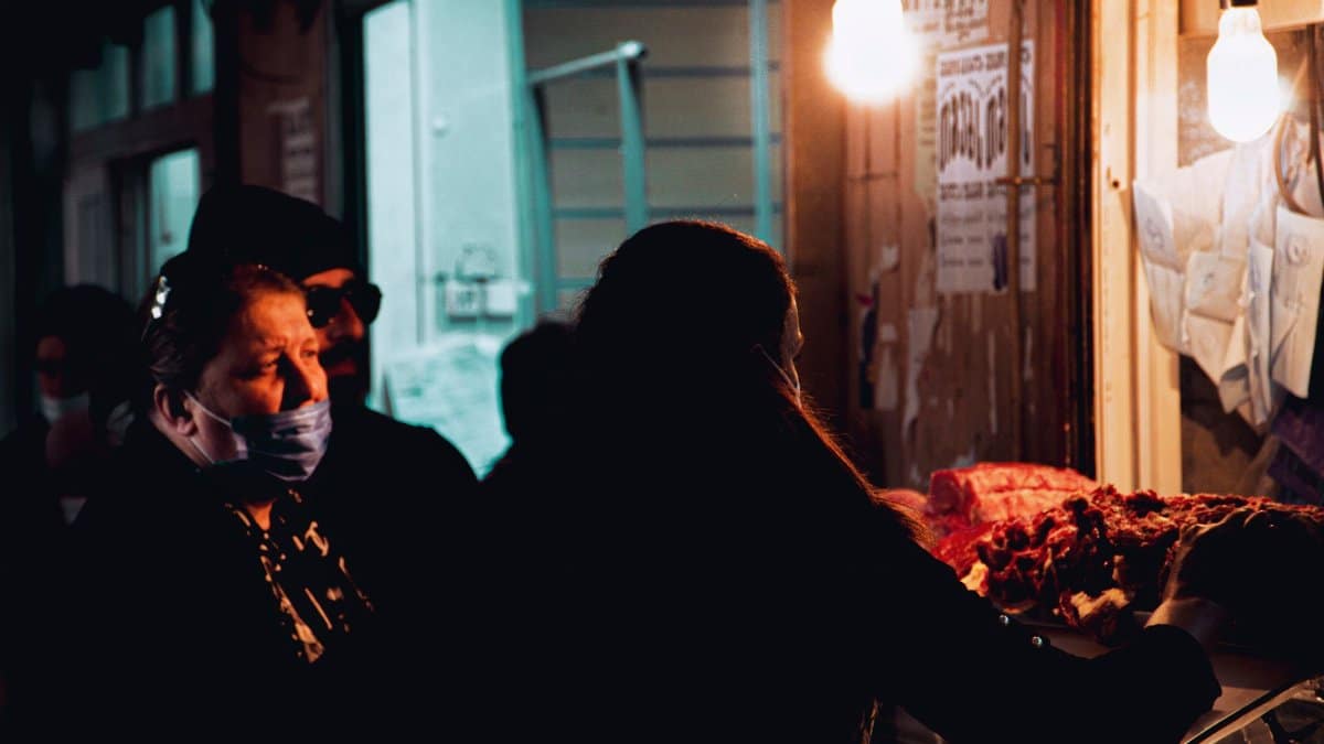A dimly lit indoor meat market stall with customers browsing fresh cuts under warm light bulbs.