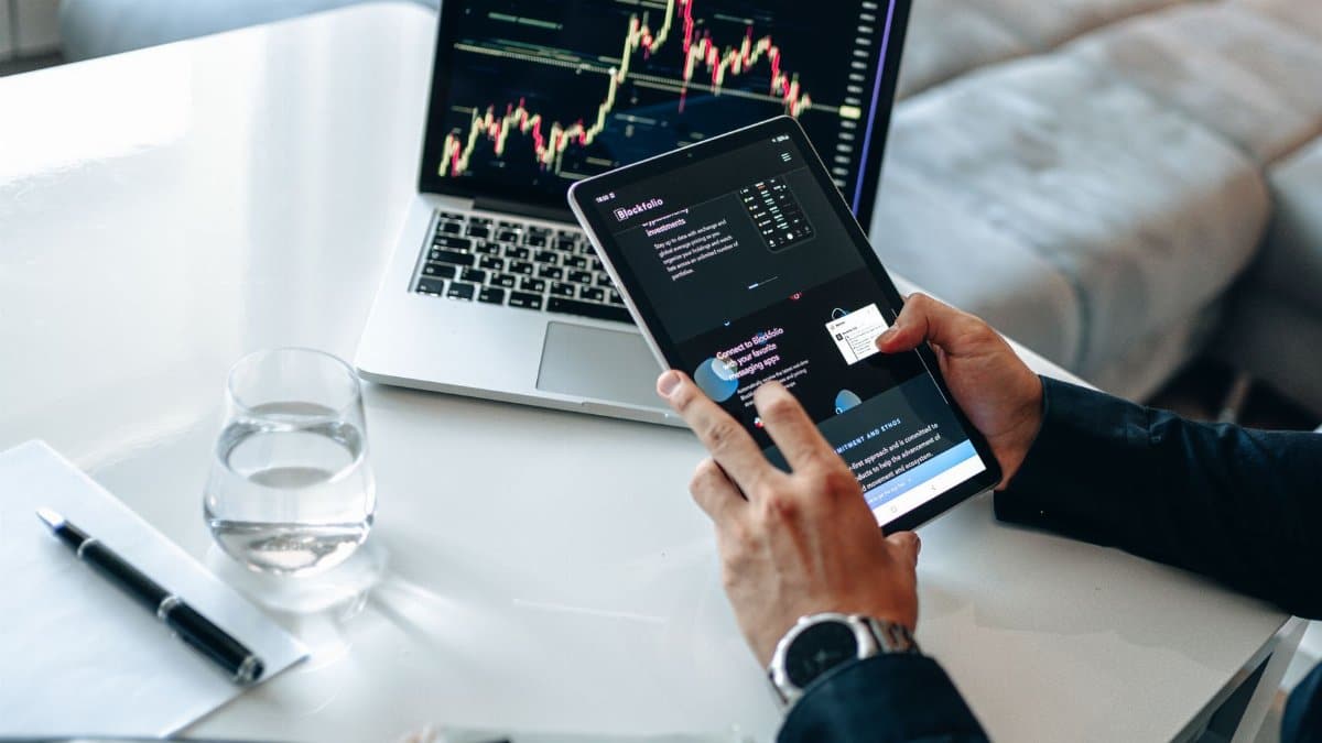 Person analyzing digital market trends on tablet with laptop showing financial charts.