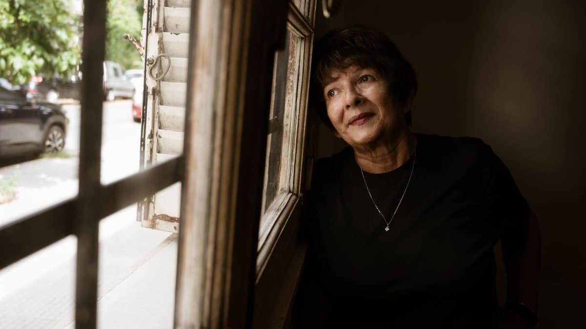 Senior woman gazing out of a window, lost in thought, with soft light illuminating her face.