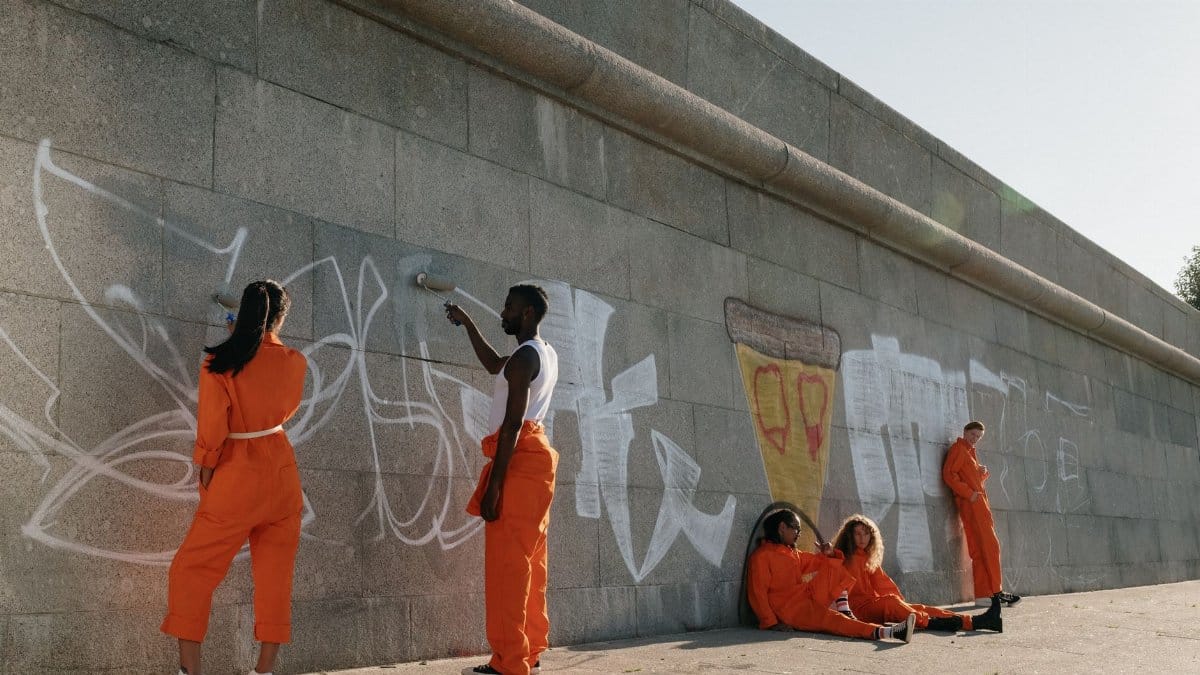 A group of people in orange outfits painting graffiti on a city wall during the day.