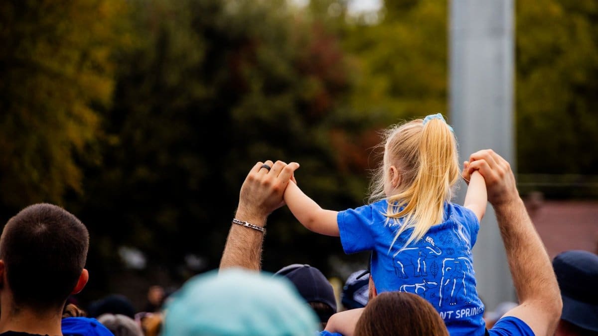 Child on shoulders in a crowd at a sunny outdoor event.