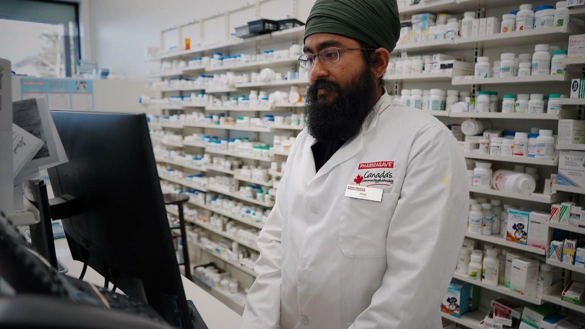Pharmacist wearing a turban working at a computer in a pharmacy store with shelves filled with medicine.