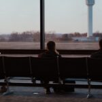 People sitting in airport terminal, watching runway scenes through large windows.