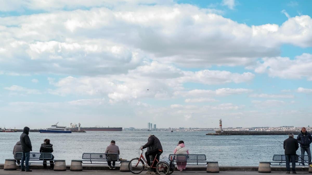 A serene seaside view with people on benches, a bicycle and distant ships under a blue cloudy sky.