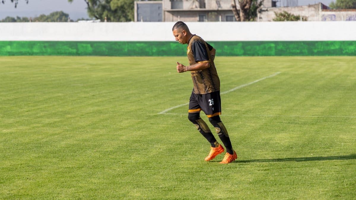 An adult male soccer player jogging on a grassy field during daytime training.