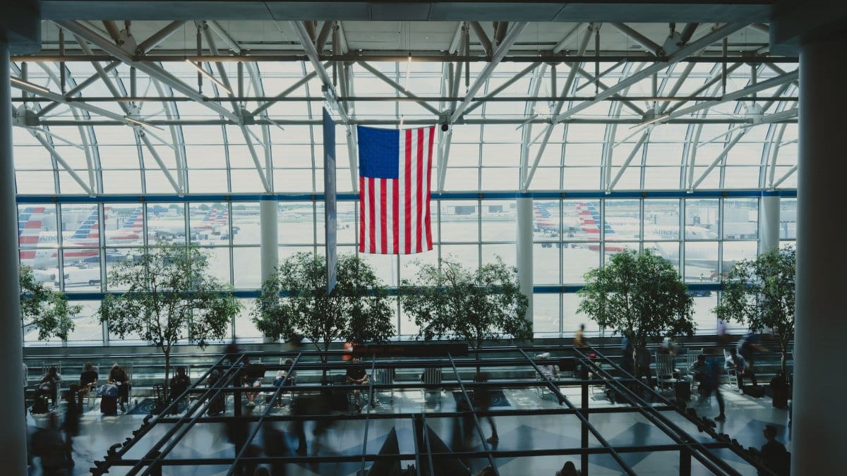 Interior view of an airport terminal's glass facade with an American flag, trees, and travelers.