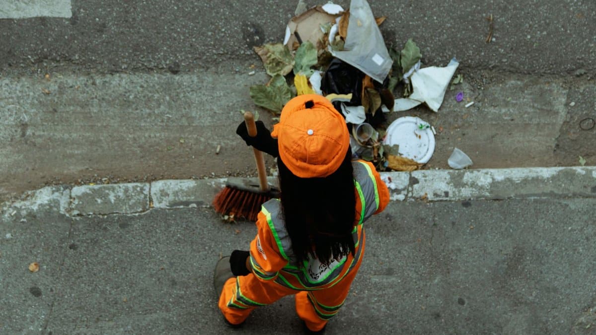 A street cleaner in an orange uniform sweeps debris from the roadside, ensuring cleanliness.