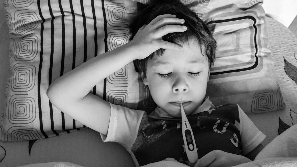 Black and white overhead view of child in casual clothing measuring body temperature with electronic thermometer while lying on bed under blanket
