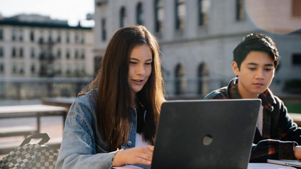 Two students collaborating on a laptop outdoors with a historic building in the background.