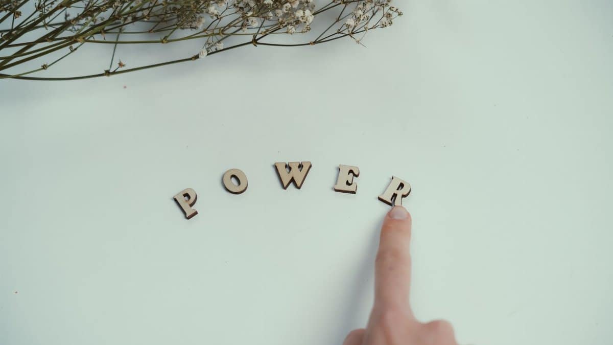 Finger points at wooden letters spelling 'Power' beside delicate flowers on a light background.