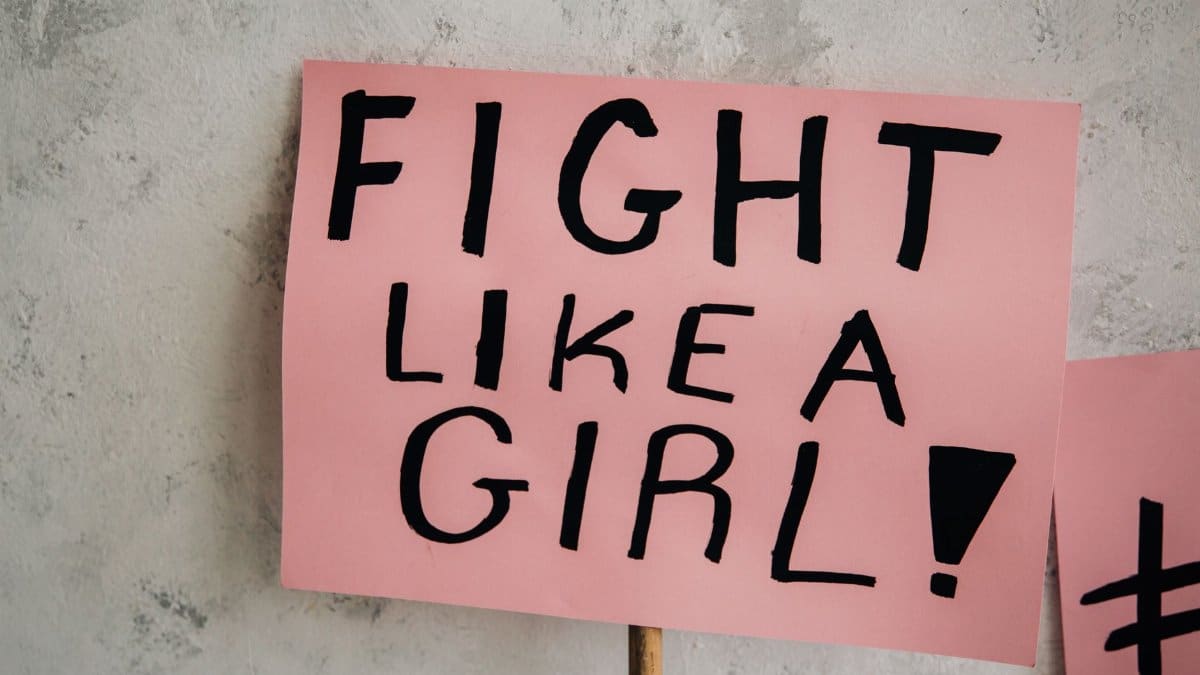 Close-up of a 'Fight Like a Girl' message on a pink placard against a gray wall.